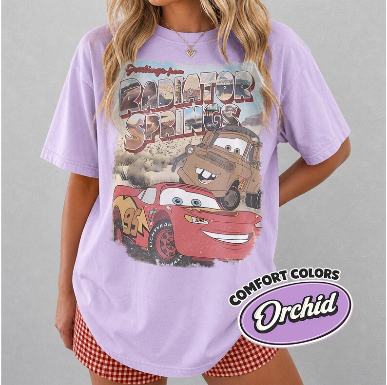 May include: A lilac-colored t-shirt featuring a graphic from the movie Cars, with Lightning McQueen and Mater. The design includes the text "Greetings from Radiator Springs." The shirt also has a "Comfort Colors" label with the word "Orchid."