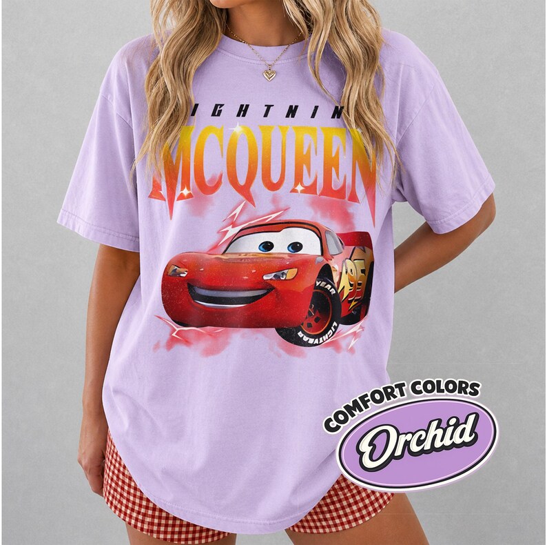 May include: A light purple t-shirt featuring a graphic of Lightning McQueen from the Cars movie. The text "LIGHTNING MCQUEEN" is printed above the car. The shirt also has the text "COMFORT COLORS" and "Orchid" in a circular design.