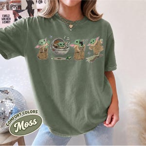 May include: A moss green short-sleeve t-shirt featuring a graphic print of a small green creature in various poses, including sitting in a floating pod, eating, and standing. The design includes small musical notes, a butterfly, and a frog.