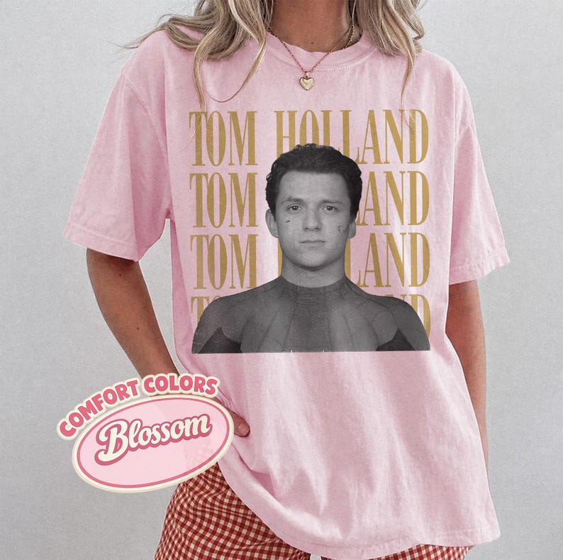 Peut inclure: T-shirt rose clair avec une image en noir et blanc de Tom Holland. Les mots "TOM HOLLAND" sont imprim&eacute;s en or verticalement &agrave; droite. Le t-shirt porte &eacute;galement les mots "COMFORT COLORS Blossom".