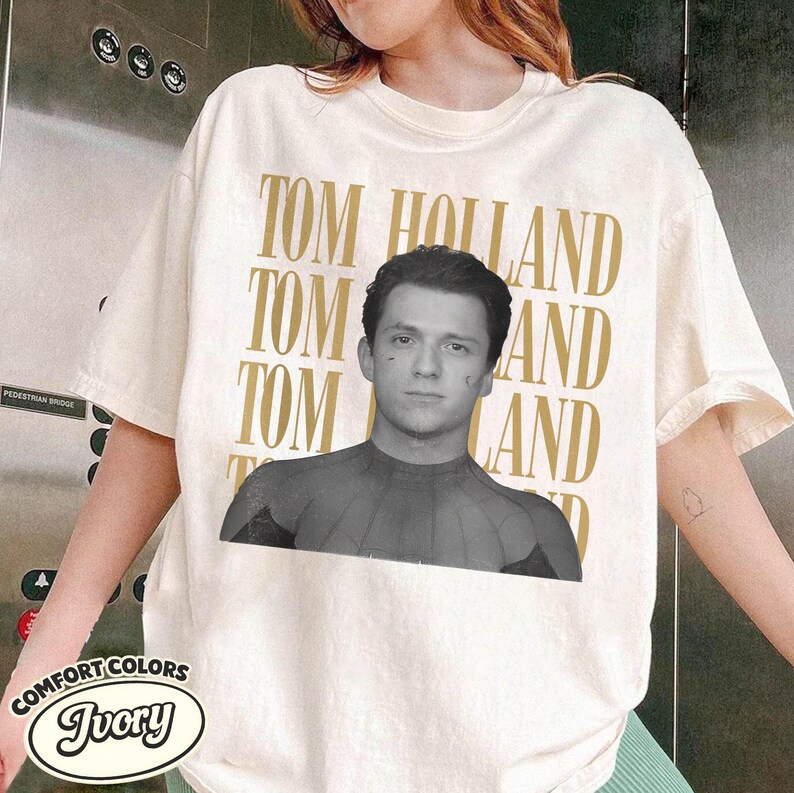 Peut inclure: T-shirt ivoire avec le nom "TOM HOLLAND" imprim&eacute; en or &agrave; plusieurs reprises. Une image en noir et blanc de Tom Holland est pr&eacute;sente sur le devant. Le t-shirt est de Comfort Colors.