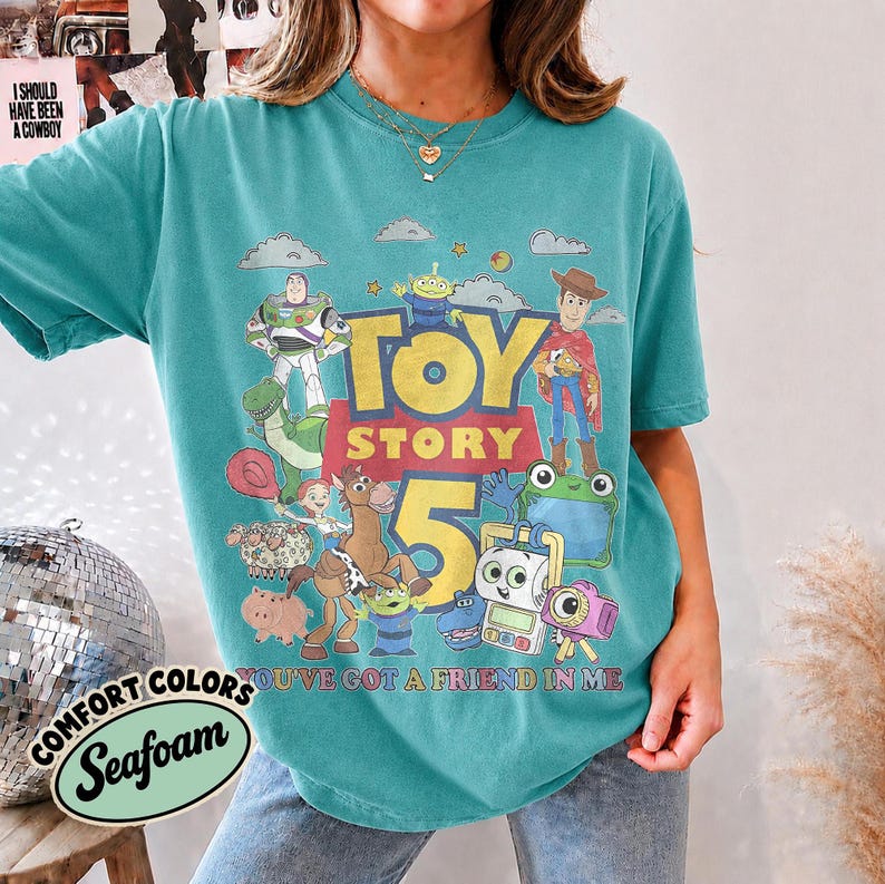 May include: A teal short-sleeve t-shirt featuring a graphic print of Toy Story characters including Buzz Lightyear, Woody, Rex, and others. The text reads "TOY STORY 5" and "YOU'VE GOT A FRIEND IN ME" in yellow and red lettering across the center chest.