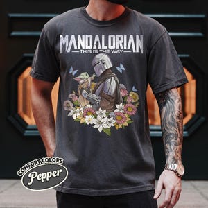May include: Dark gray t-shirt featuring the text "MANDALORIAN - THIS IS THE WAY" above an illustration of the Mandalorian and Grogu surrounded by flowers and butterflies. The shirt has a Comfort Colors Pepper label.