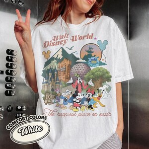 Pu&ograve; includere: T-shirt bianco con una grafica colorata di Walt Disney World con monumenti e personaggi iconici. Il design include il testo "Walt Disney World" e "The happiest place on earth". La maglietta &egrave; di Comfort Colors.
