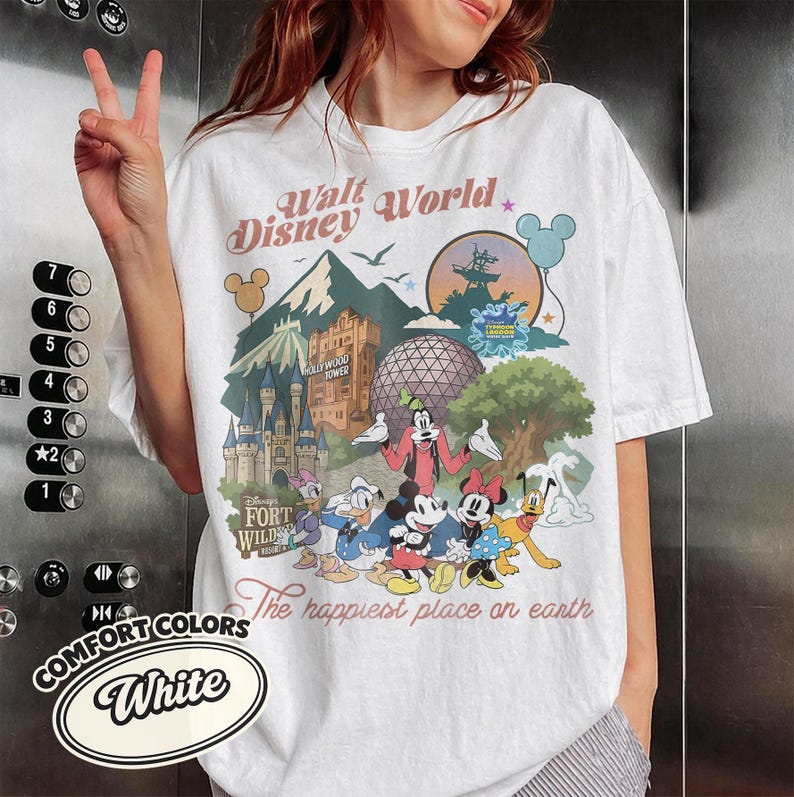 Pu&ograve; includere: T-shirt bianco con una grafica colorata di Walt Disney World con monumenti e personaggi iconici. Il design include il testo "Walt Disney World" e "The happiest place on earth". La maglietta &egrave; di Comfort Colors.