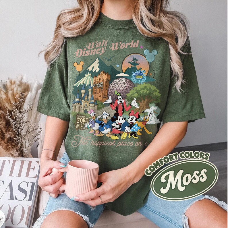 Pu&ograve; includere: T-shirt verde con una grafica colorata di personaggi e monumenti di Disney World. La maglietta presenta la scritta "Walt Disney World" e "The happiest place on earth". La maglietta &egrave; etichettata "Comfort Colors Moss".
