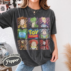 May include: A charcoal grey short-sleeve t-shirt featuring a grid of Toy Story characters including Woody, Rex, Bo Peep, Slinky Dog, Jessie, Buzz Lightyear, and Forky, with the text Toy Story in the center and a small green tablet reading Hi.