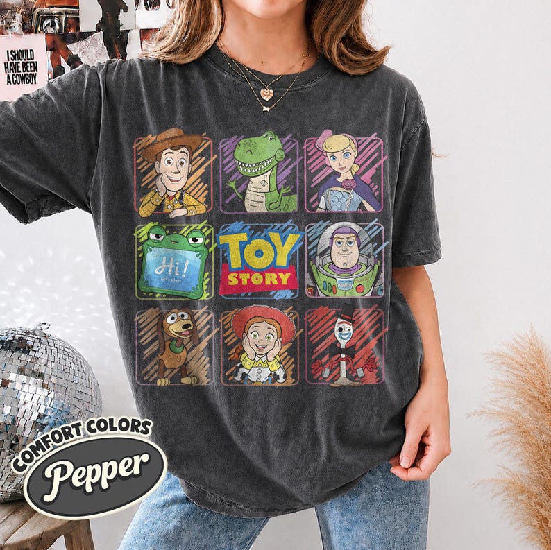May include: A charcoal grey short-sleeve t-shirt featuring a grid of Toy Story characters including Woody, Rex, Bo Peep, Slinky Dog, Jessie, Buzz Lightyear, and Forky, with the text Toy Story in the center and a small green tablet reading Hi.