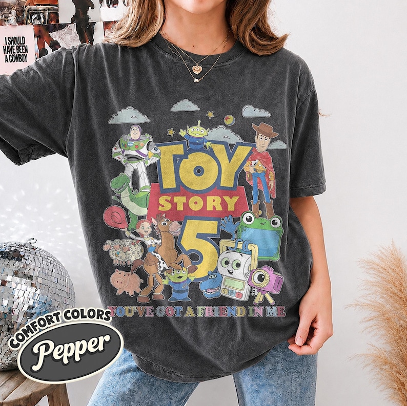 May include: A charcoal gray short-sleeve t-shirt featuring a graphic print of Toy Story characters including Woody, Buzz Lightyear, and Jessie. The text reads Toy Story 5 and You've Got a Friend in Me. The shirt has a vintage-style washed appearance.
