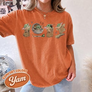 May include: An orange short-sleeve t-shirt featuring a graphic print of a small green creature in various poses, including sitting in a floating pod, holding a cup, and standing, accompanied by a small green frog and musical notes.