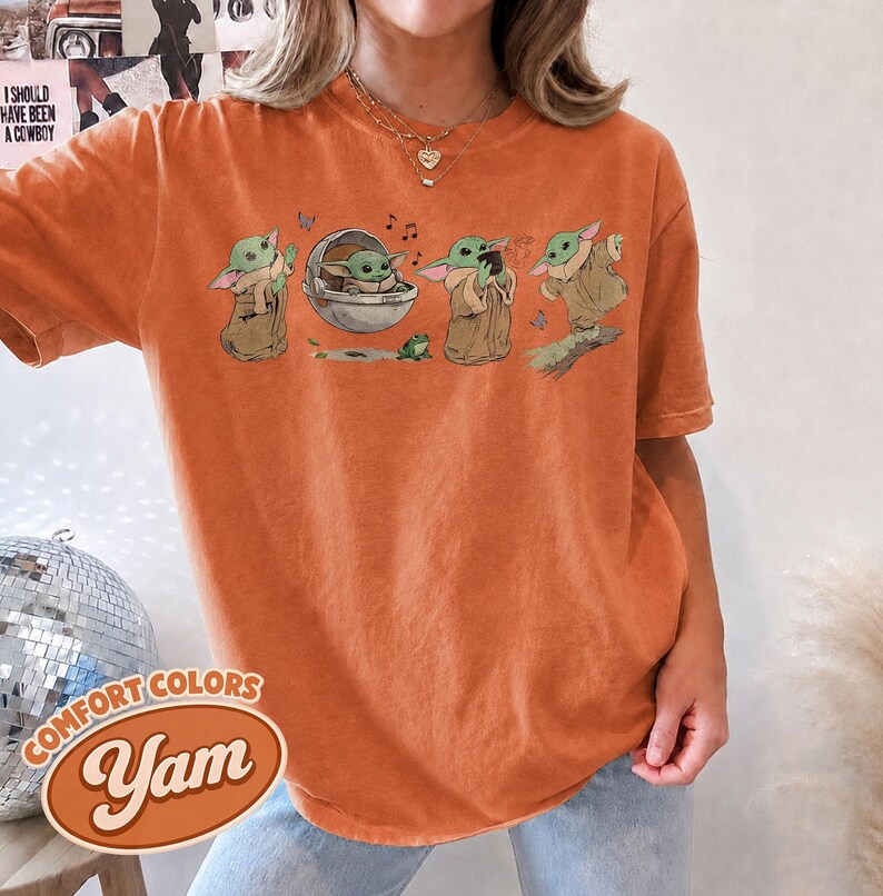May include: An orange short-sleeve t-shirt featuring a graphic print of a small green creature in various poses, including sitting in a floating pod, holding a cup, and standing, accompanied by a small green frog and musical notes.