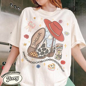 Peut inclure: Un t-shirt ivoire avec une impression graphique repr&eacute;sentant une botte de cow-boy &agrave; motif vache, un chapeau rouge et des blocs &eacute;pelant TS. Le design inclut le nom Taylor, le chiffre 13, un n&oelig;ud, des fleurs et une perle avec le texte I KNEW IT.