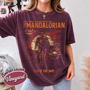 May include: A burgundy short-sleeve t-shirt featuring a graphic of The Mandalorian and Grogu. The text reads: BOUNTY HUNTING IS A COMPLICATED PROFESSION... THE MANDALORIAN THIS IS THE WAY. The shirt has a distressed, vintage-style wash finish.