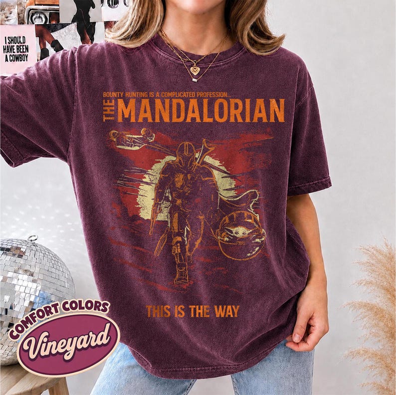 May include: A burgundy short-sleeve t-shirt featuring a graphic of The Mandalorian and Grogu. The text reads: BOUNTY HUNTING IS A COMPLICATED PROFESSION... THE MANDALORIAN THIS IS THE WAY. The shirt has a distressed, vintage-style wash finish.