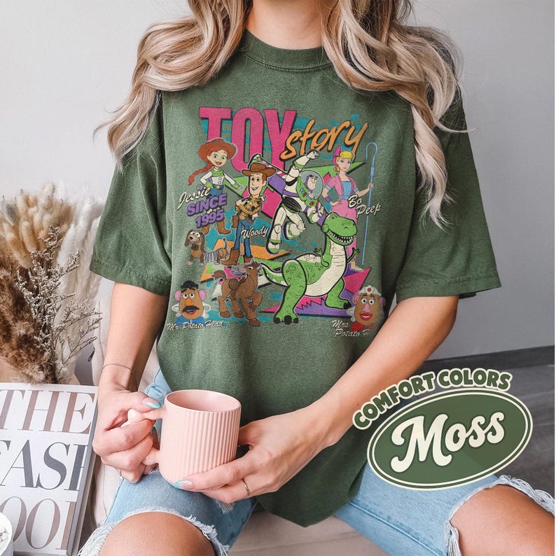 May include: An olive green t-shirt featuring a graphic of Toy Story characters, including Woody, Buzz Lightyear, and Jessie. The design includes the text "Toy Story" and "Since 1995". The shirt is part of the "Comfort Colors" collection and is in the color "Moss".