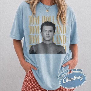 Peut inclure: T-shirt bleu clair avec une image en noir et blanc de Tom Holland et le texte "TOM HOLLAND" en or. Le t-shirt a des manches courtes et une coupe d&eacute;contract&eacute;e. Le t-shirt porte &eacute;galement le texte "COMFORT COLORS Chambray" dans un ovale bleu.