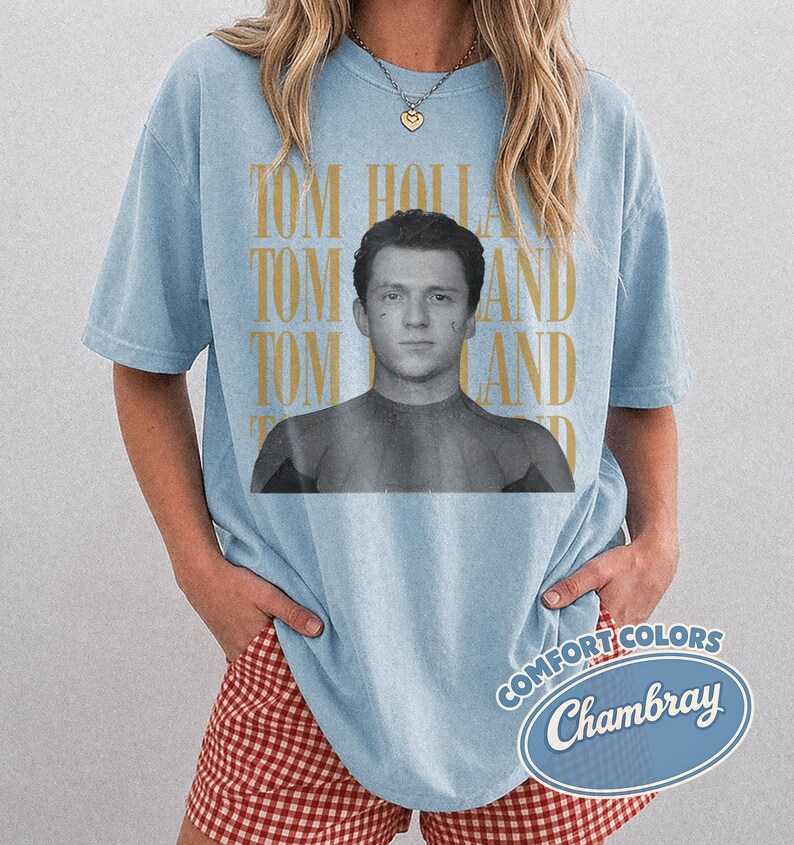 Peut inclure: T-shirt bleu clair avec une image en noir et blanc de Tom Holland et le texte "TOM HOLLAND" en or. Le t-shirt a des manches courtes et une coupe d&eacute;contract&eacute;e. Le t-shirt porte &eacute;galement le texte "COMFORT COLORS Chambray" dans un ovale bleu.