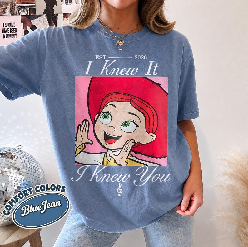 May include: A blue jean colored t-shirt featuring a graphic of a cartoon character with red hair and a red hat. The text "EST 2026", "I Knew It", and "I Knew You" with a musical note symbol are printed in white above and below the central graphic.