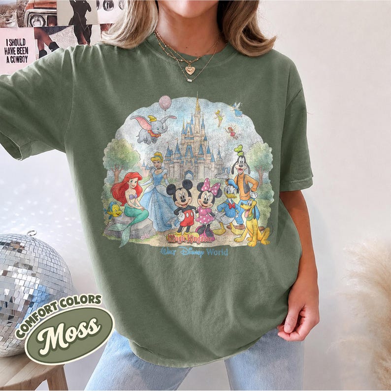 Op de afbeelding: Een mosgroen t-shirt met korte mouwen en een grafische print van een kasteel en diverse tekenfilmfiguren, waaronder Mickey Mouse, Minnie Mouse, Donald Duck, Goofy, Pluto, Ari&euml;l en Dumbo. De tekst luidt Magic Kingdom en Walt Disney World.