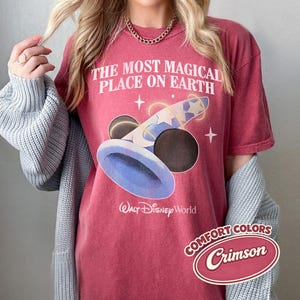 K&ouml;nnte beinhalten: Karmesinrotes T-Shirt mit dem Text "THE MOST MAGICAL PLACE ON EARTH" und einer Grafik von Mickey Mouses Zaubererhut. Das Walt Disney World-Logo befindet sich unter der Grafik. Das Shirt hat auch ein "Comfort Colors"-Etikett.