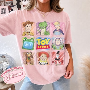 May include: A light pink t-shirt featuring a grid of Toy Story characters including Woody, Rex, Bo Peep, a green frog with the text Hi! Let's play!, Buzz Lightyear, Slinky Dog, Jessie, and Forky, with the Toy Story logo in the center.