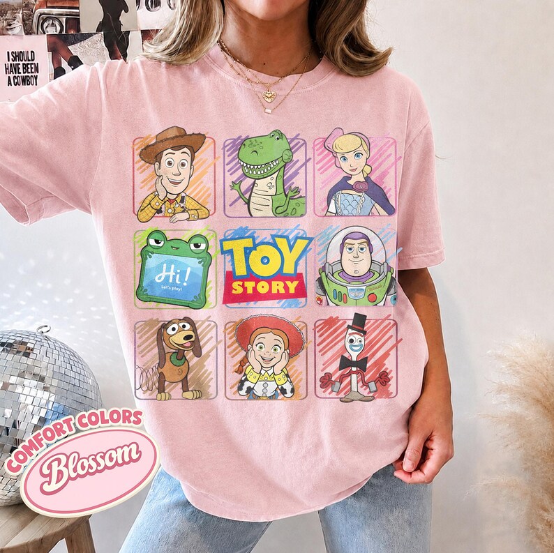 May include: A light pink t-shirt featuring a grid of Toy Story characters including Woody, Rex, Bo Peep, a green frog with the text Hi! Let's play!, Buzz Lightyear, Slinky Dog, Jessie, and Forky, with the Toy Story logo in the center.