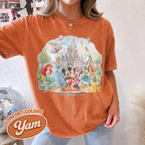 Op de afbeelding: Een kort mouwloos t-shirt in gebrand oranje met een grafische print van Disney-figuren waaronder Mickey Mouse, Minnie Mouse, Ariel, Assepoester, Dumbo, Donald Duck, Goofy en Pluto voor een kasteel met de tekst Magic Kingdom Walt Disney World.