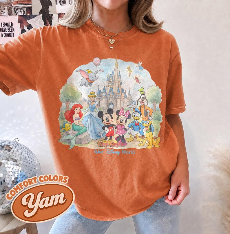 Op de afbeelding: Een kort mouwloos t-shirt in gebrand oranje met een grafische print van Disney-figuren waaronder Mickey Mouse, Minnie Mouse, Ariel, Assepoester, Dumbo, Donald Duck, Goofy en Pluto voor een kasteel met de tekst Magic Kingdom Walt Disney World.