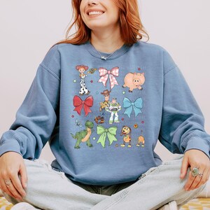 May include: A dusty blue crewneck sweatshirt featuring a graphic print of various cartoon characters and colorful bows. The design includes a cowgirl, a cowboy, a space ranger, a green dinosaur, a pig, and a dog, interspersed with red, pink, blue, and green bows.