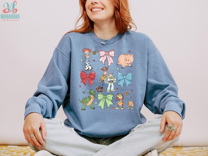 May include: A dusty blue crewneck sweatshirt featuring a graphic print of various cartoon characters and colorful bows. The design includes a cowgirl, a cowboy, a space ranger, a green dinosaur, a pig, and a dog, interspersed with red, pink, blue, and green bows.