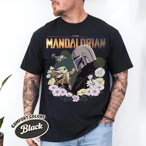 May include: Black t-shirt featuring a graphic of The Mandalorian and Grogu surrounded by flowers. The text "THE MANDALORIAN" is displayed above the characters. The shirt has a Comfort Colors label.