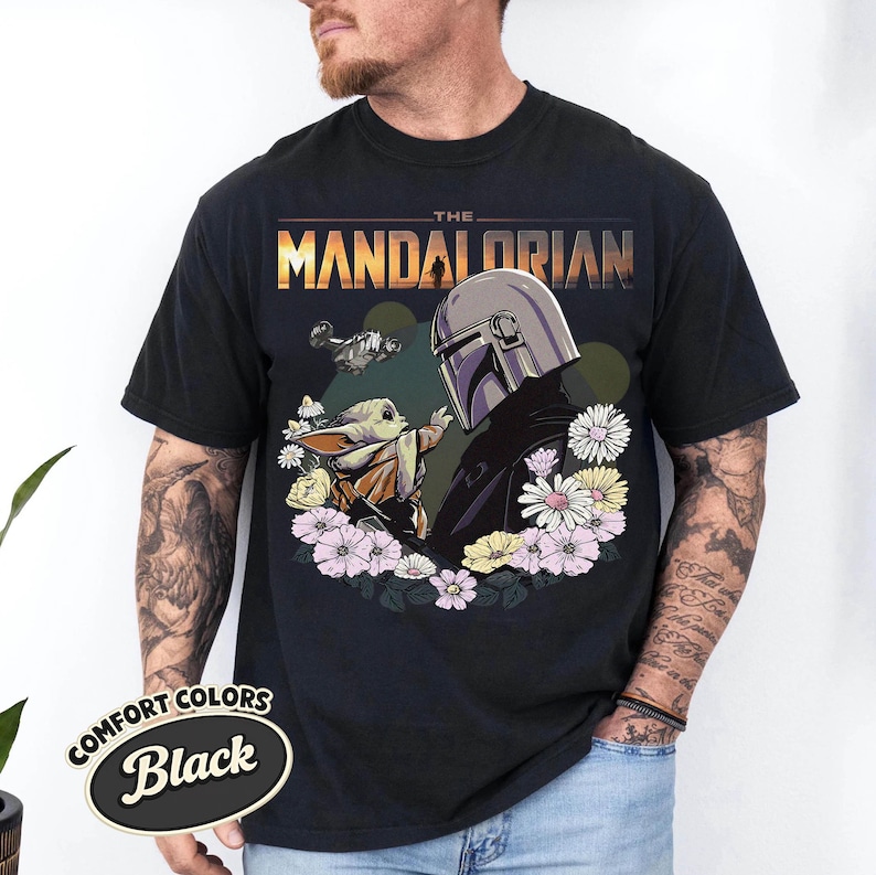 May include: Black t-shirt featuring a graphic of The Mandalorian and Grogu surrounded by flowers. The text "THE MANDALORIAN" is displayed above the characters. The shirt has a Comfort Colors label.
