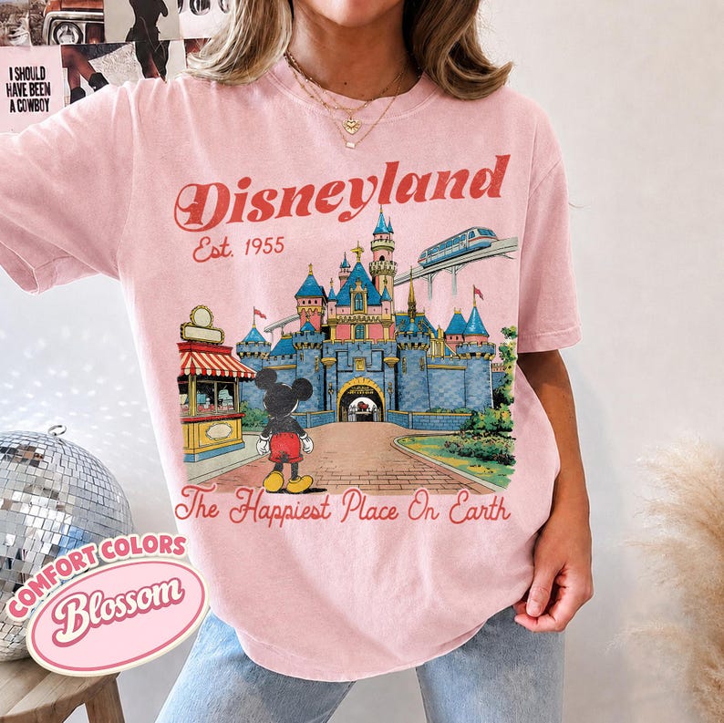 May include: A light pink t-shirt featuring a graphic of the Disneyland castle, Mickey Mouse, and a monorail. The text reads Disneyland Est. 1955 and The Happiest Place On Earth. The shirt has a crew neckline and short sleeves with a relaxed fit.