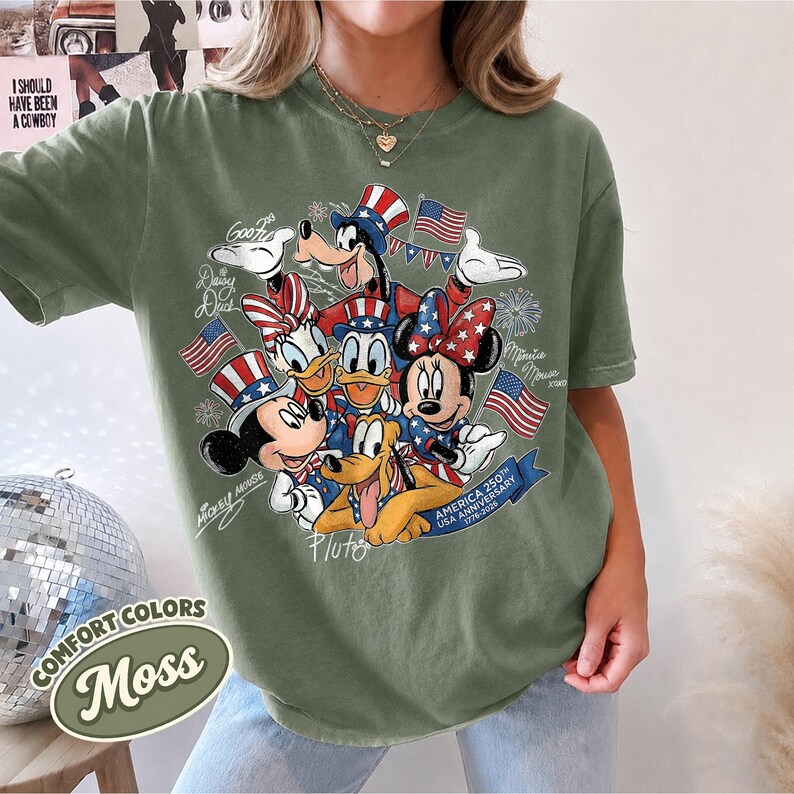 May include: Moss green t-shirt featuring a patriotic design with Disney characters Mickey Mouse, Minnie Mouse, Donald Duck, Daisy Duck, Goofy, and Pluto. The design includes American flags and text that reads "America 250th Anniversary USA 1776-2026". The shirt also has the text "Comfort Colors Moss".