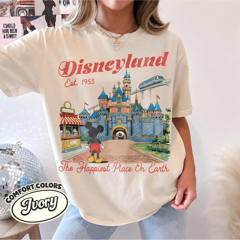 May include: An ivory short-sleeve t-shirt featuring a graphic print of the Disneyland castle, Mickey Mouse, and a monorail. The text reads Disneyland, Est. 1955, and The Happiest Place On Earth in a vintage-style red font on the front of the shirt.