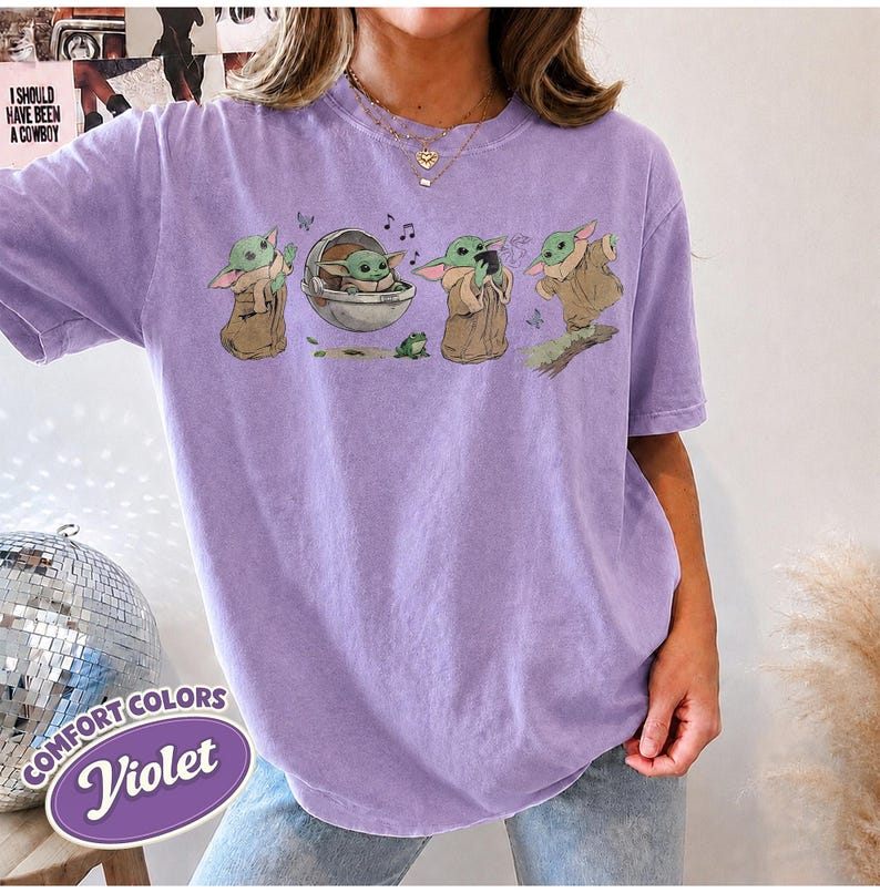 May include: A violet short-sleeved t-shirt featuring a graphic print of a green alien character in various poses, including sitting in a floating pod, standing, and holding a cup. The design includes small musical notes and a frog illustration.