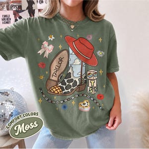 Peut inclure: Un t-shirt vert mousse avec une impression graphique d'une botte de cow-boy &agrave; motifs de vache, un chapeau rouge, le nom TAYLOR, le chiffre 13, des blocs empil&eacute;s avec les lettres T, S et un collier de perles avec le texte I KNEW IT I KNEW YOU.