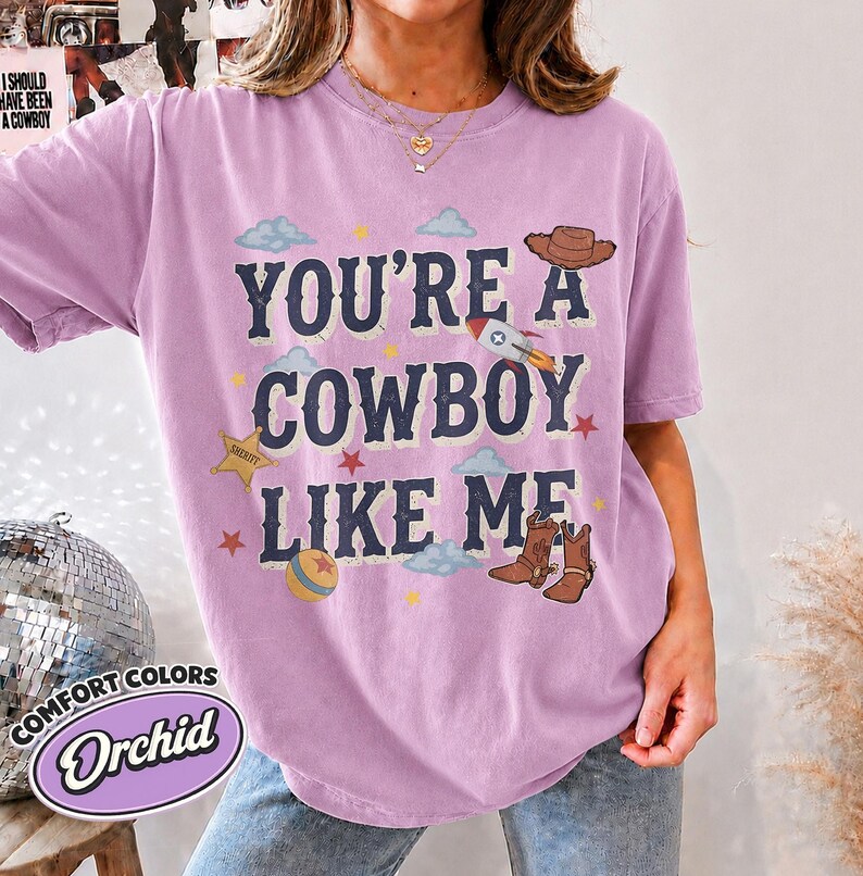 May include: An orchid-colored short-sleeve t-shirt featuring the text "YOU'RE A COWBOY LIKE ME" in dark blue letters. The graphic includes a brown cowboy hat, a rocket ship, a gold sheriff star, a colorful ball, and brown cowboy boots.