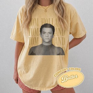 Peut inclure: T-shirt jaune beurre avec une image en noir et blanc de Tom Holland. Les mots "TOM HOLLAND" sont imprim&eacute;s verticalement sur le c&ocirc;t&eacute; gauche. Les mots "COMFORT COLORS" et "Butter" sont imprim&eacute;s sur un ovale jaune.