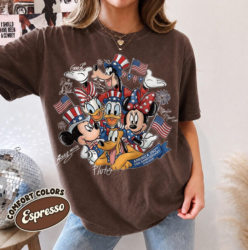 May include: Espresso-colored t-shirt featuring a graphic of Disney characters in patriotic attire, including Mickey and Minnie Mouse, Donald Duck, and Pluto, with American flags. The shirt has the text "America 250th USA Anniversary 1776-2026" and "Comfort Colors Espresso".