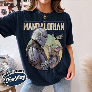 Puede incluir: Una camiseta azul marino oscuro con un estampado gr&aacute;fico de The Mandalorian y Grogu dentro de un marco circular. El texto THE MANDALORIAN est&aacute; impreso en una fuente negrita y clara sobre los personajes, con una peque&ntilde;a silueta de nave espacial.