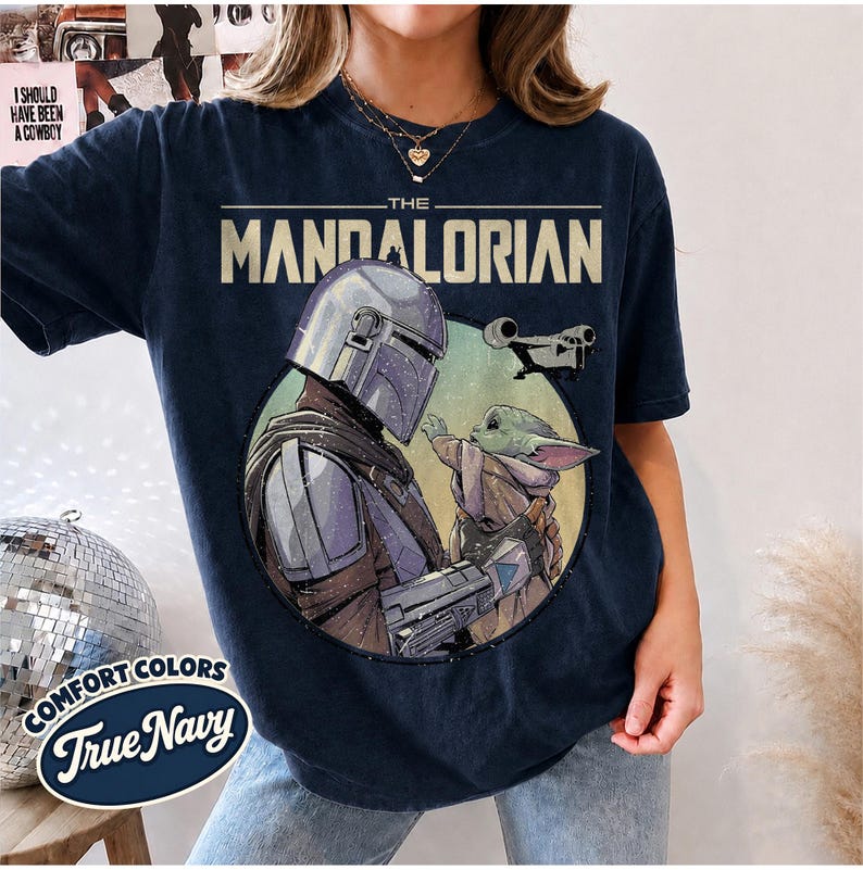 Puede incluir: Una camiseta azul marino oscuro con un estampado gr&aacute;fico de The Mandalorian y Grogu dentro de un marco circular. El texto THE MANDALORIAN est&aacute; impreso en una fuente negrita y clara sobre los personajes, con una peque&ntilde;a silueta de nave espacial.