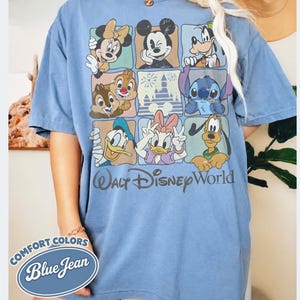 May include: Blue short-sleeve t-shirt featuring a grid of Disney characters including Mickey Mouse, Minnie Mouse, Donald Duck, and Stitch. The shirt has the words "Walt Disney World" printed on it. The shirt is labeled "Comfort Colors Blue Jean".