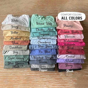 May include: Stacks of folded t-shirts in various colors, including white, blue, pink, and green. Each stack is labeled with the color name, such as "White," "Peachy," and "Blue Jean." The text "COMFORT COLORS ALL COLORS" is displayed at the top.
