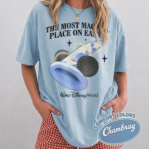K&ouml;nnte beinhalten: Hellblaues T-Shirt mit einer Grafik eines blau-wei&szlig;en Zauberhuts und schwarzen Mickey Mouse-Ohren. Der Text "THE MOST MAGICAL PLACE ON EARTH" und "Walt Disney World" sind auf dem Shirt aufgedruckt.