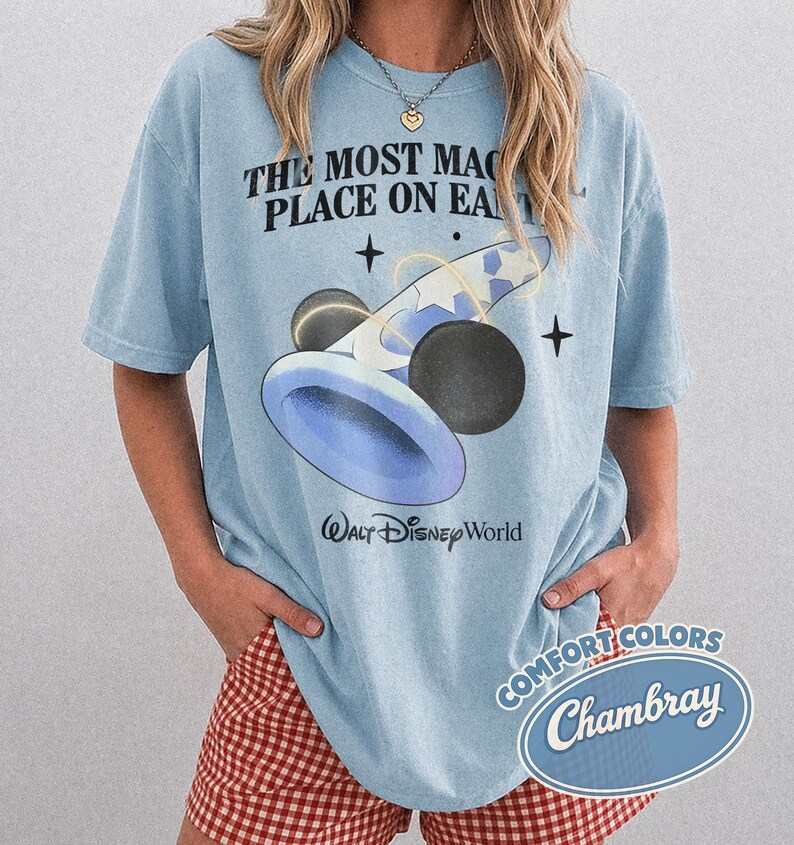 K&ouml;nnte beinhalten: Hellblaues T-Shirt mit einer Grafik eines blau-wei&szlig;en Zauberhuts und schwarzen Mickey Mouse-Ohren. Der Text "THE MOST MAGICAL PLACE ON EARTH" und "Walt Disney World" sind auf dem Shirt aufgedruckt.