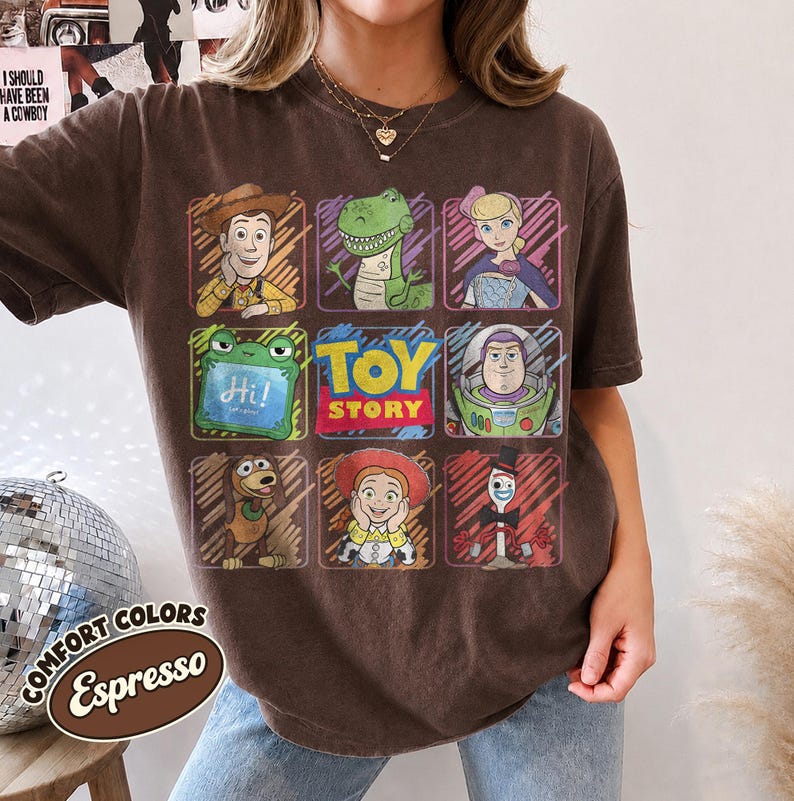May include: A dark brown short-sleeve t-shirt featuring a grid of Toy Story character illustrations including Woody, Rex, Bo Peep, Slinky Dog, Jessie, Forky, and Buzz Lightyear, with the text TOY STORY centered in a red and yellow box.