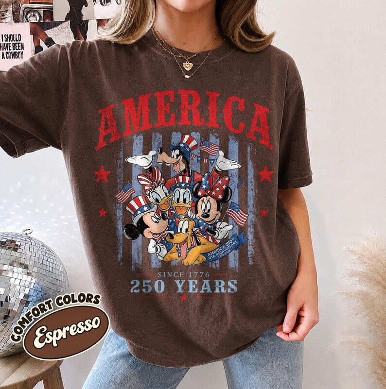 May include: A brown short-sleeve t-shirt featuring a graphic of cartoon characters wearing patriotic hats. The text reads AMERICA, SINCE 1776, 250 YEARS, and AMERICA 250TH USA ANNIVERSARY. The design includes red stars and blue vertical stripes.