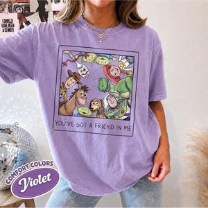 Puede incluir: Una camiseta violeta Comfort Colors con un gr&aacute;fico enmarcado de personajes de Toy Story. El gr&aacute;fico incluye a Woody, Buzz Lightyear y otros personajes, con el texto "YOU'VE GOT A FRIEND IN ME".