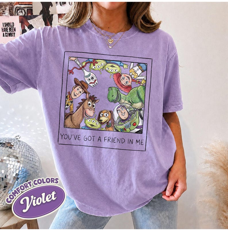 Puede incluir: Una camiseta violeta Comfort Colors con un gr&aacute;fico enmarcado de personajes de Toy Story. El gr&aacute;fico incluye a Woody, Buzz Lightyear y otros personajes, con el texto "YOU'VE GOT A FRIEND IN ME".